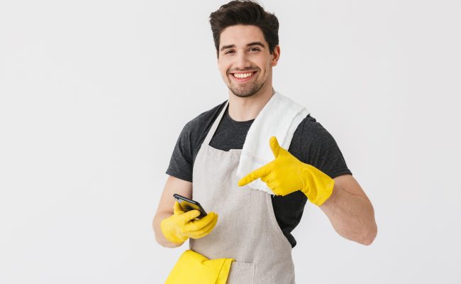 Handsome brunette houseman wearing apron standing isolated over white background, showing mobile phone